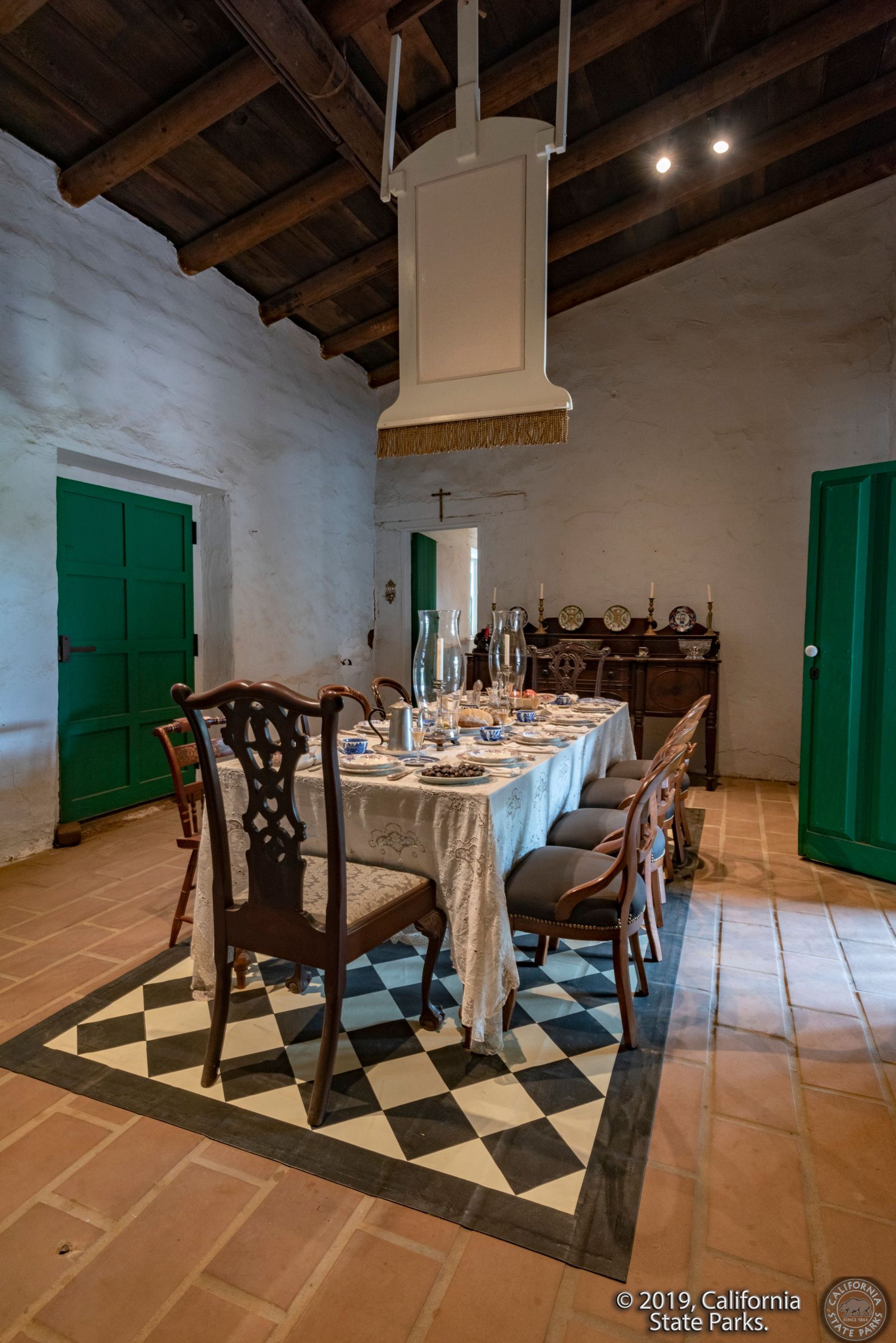 Photo of the Dining Room in La Casa de Estudillo. There is a dining room table set up for a nice meal and there is a large "shoofly" attached to the ceiling.