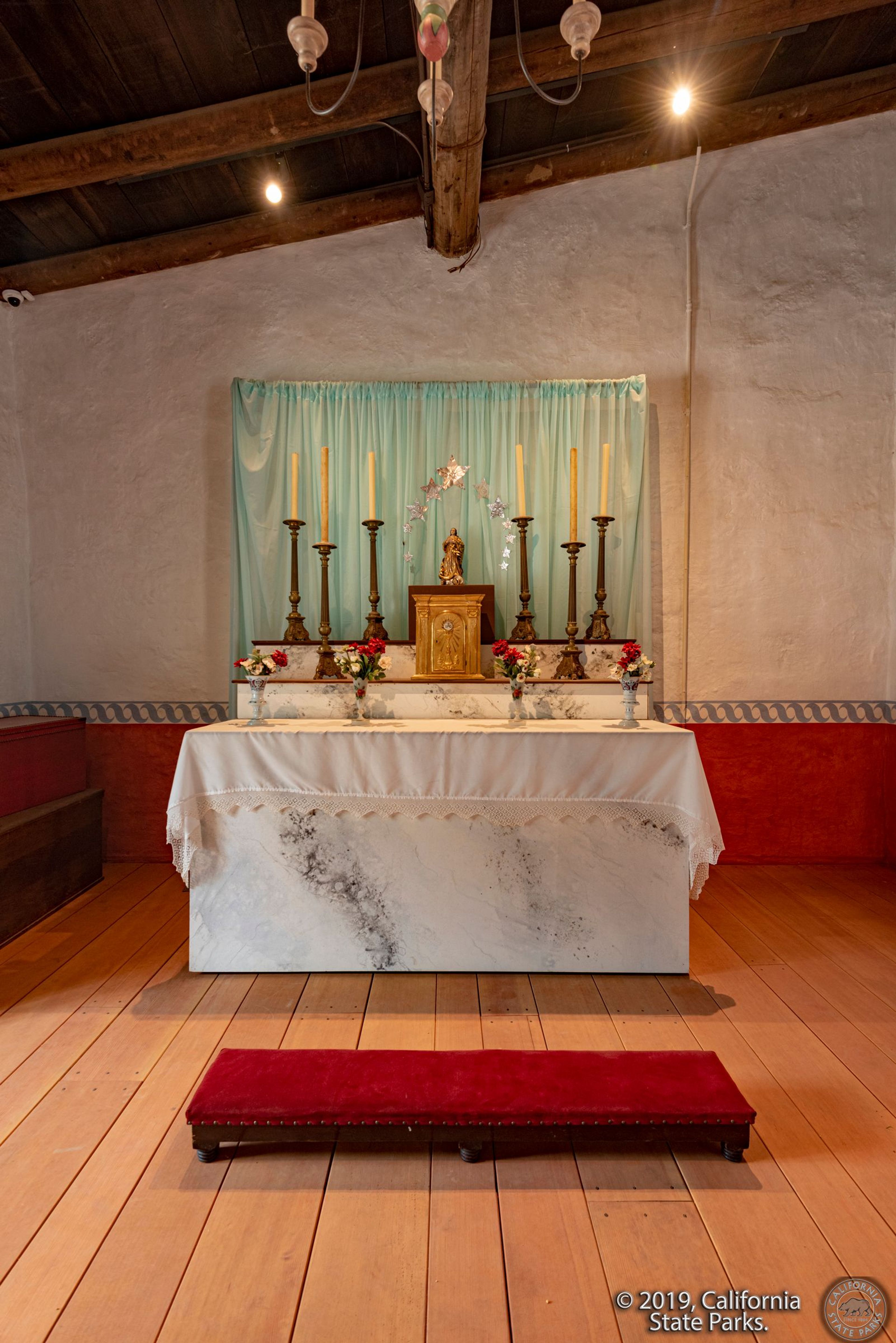 Photo of the altar set up in the sala of La Casa de Estudillo. There is a sheer curtain with tin stars attached to it behind the altar. There is a kneeler in the foreground.