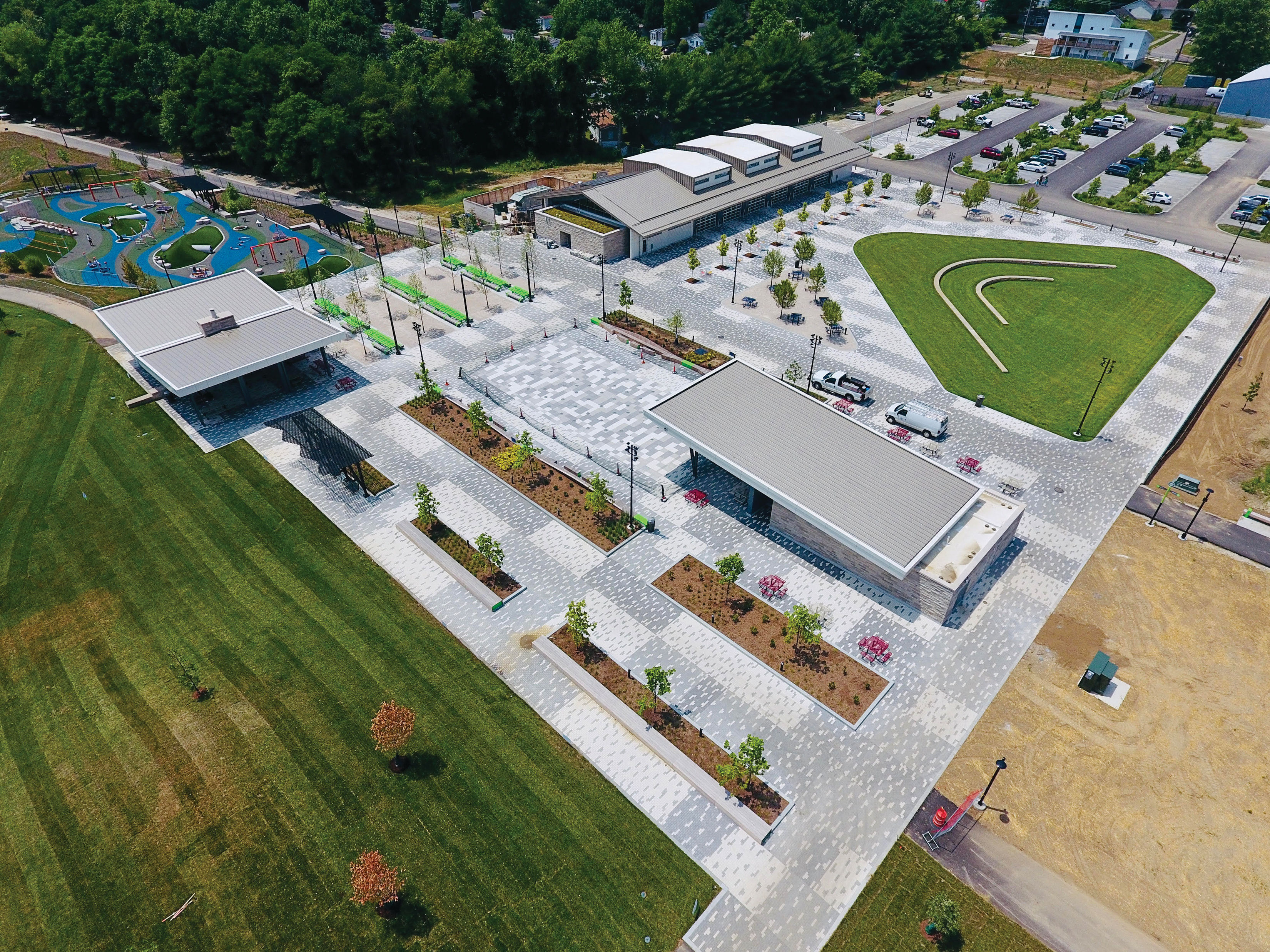 A newly inaugurated urban park along a river, with a modern playground and a multi-sport court, bustling with families.
