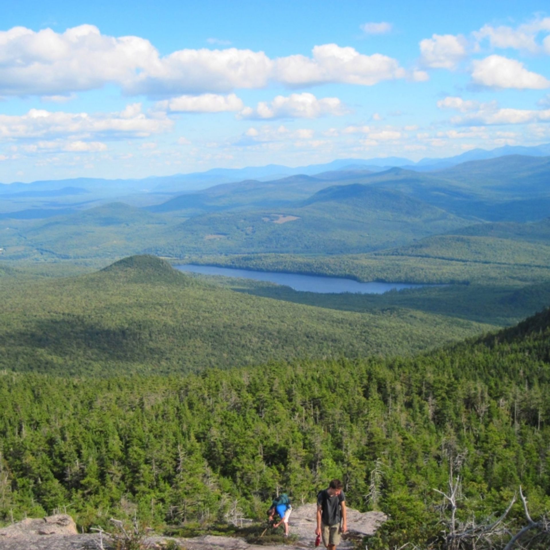 A view of the mountains and lake behind two hikers climbing.