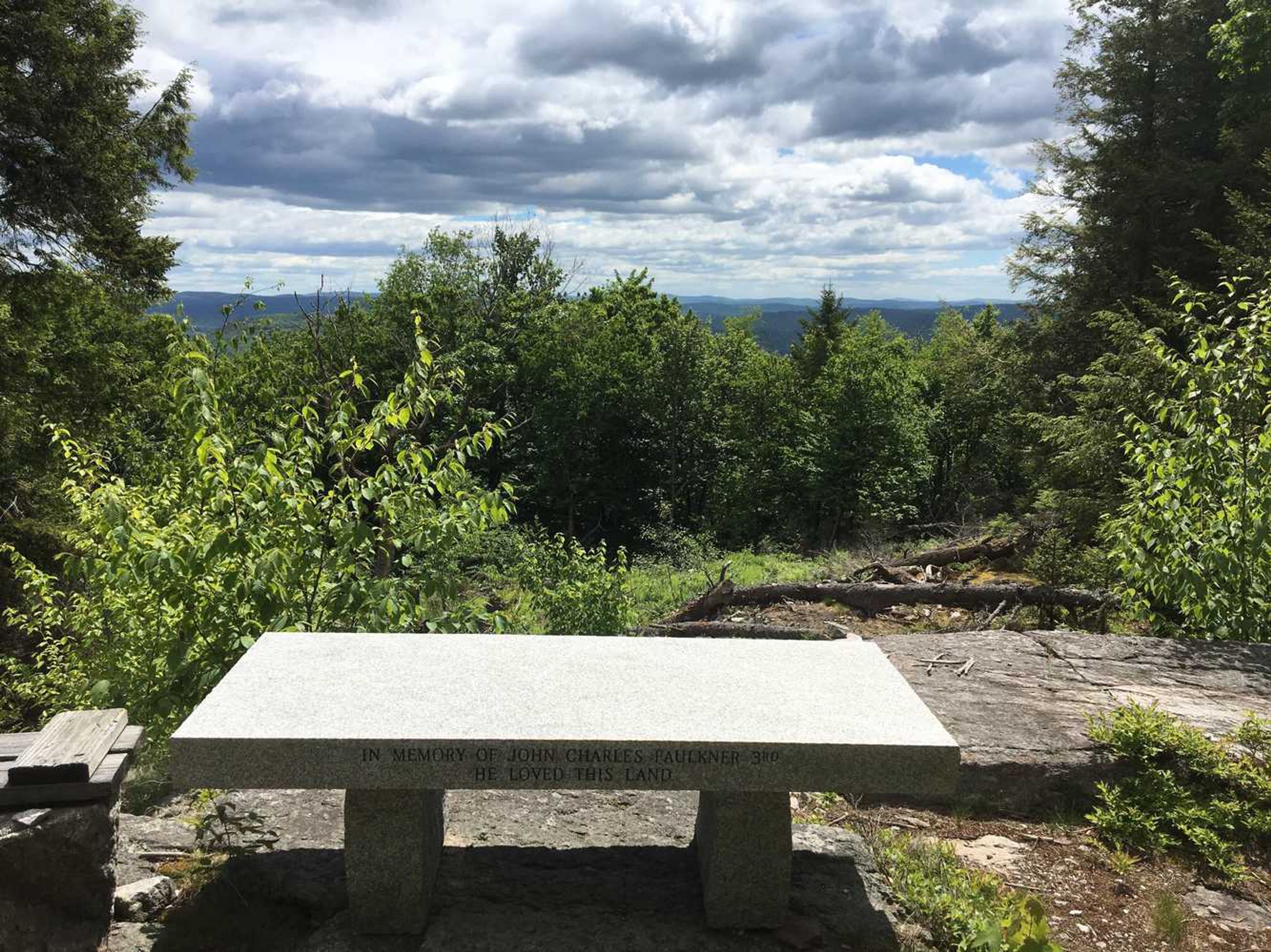 A stone bench that reads, "In memory of John Charles Faulkner 3rd. He loved this land," with the mountains beyond.