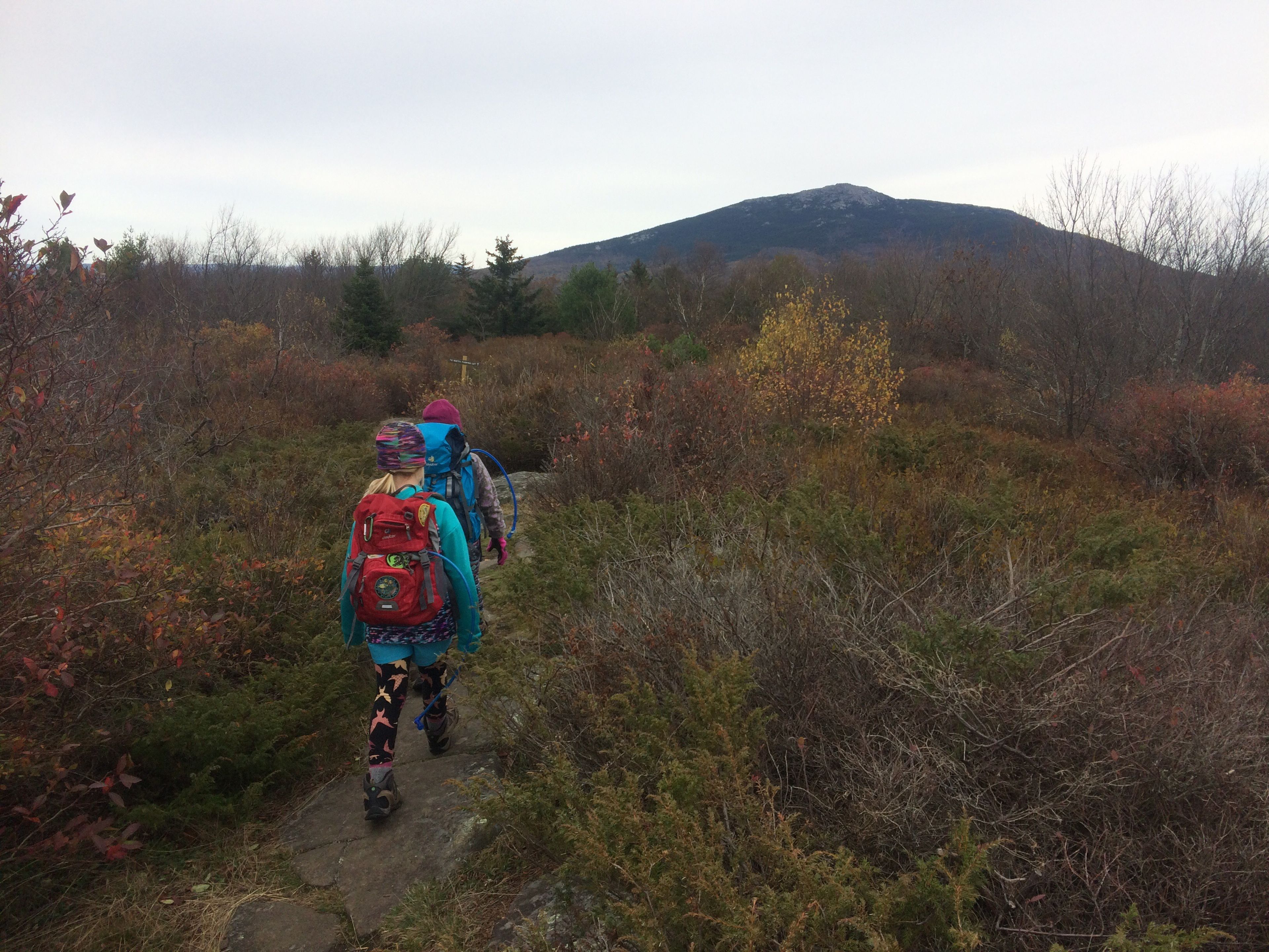 A daughter walks behind her mother on a trail looking toward Mount Monadnock.