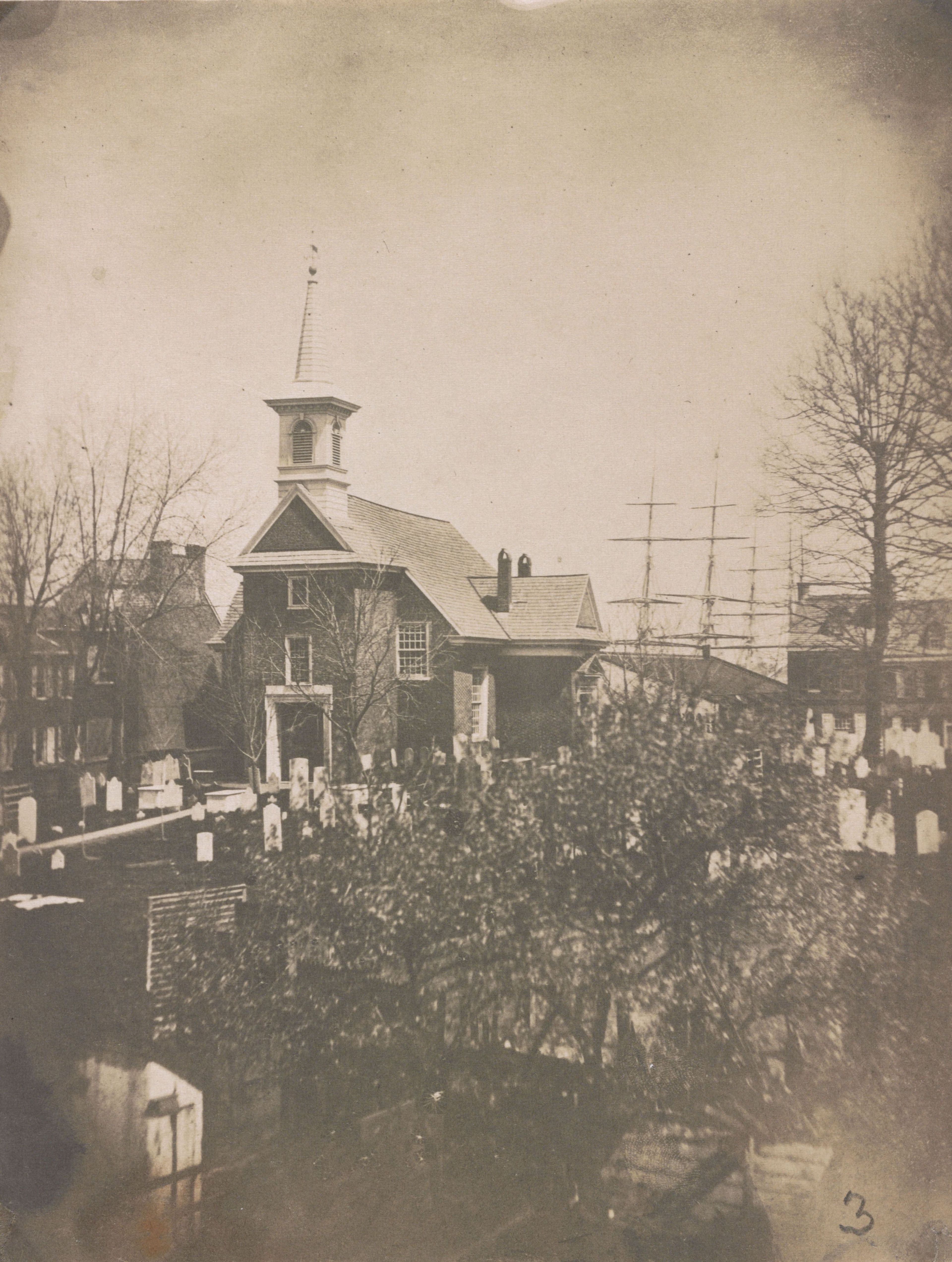 This 1850s photograph shows a bird's-eye view of the Gloria Dei Church, with the cemetery in the foreground and the masts of ships visible in the background.