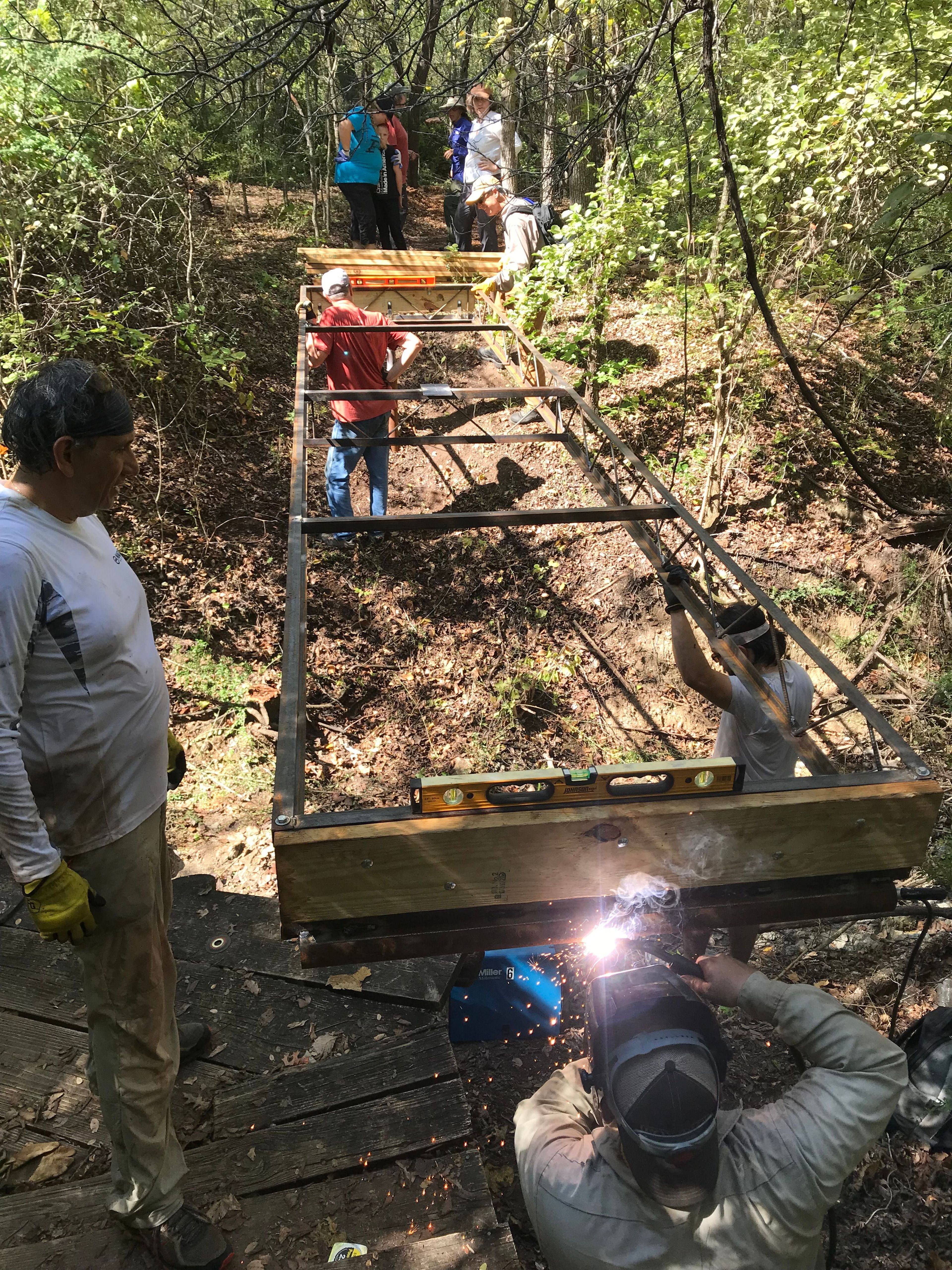 Daniel Price Natural Resource Manager welds the frame of the bridge in place while volunteers hold the frame steady. Photo by FWNCR Staff.