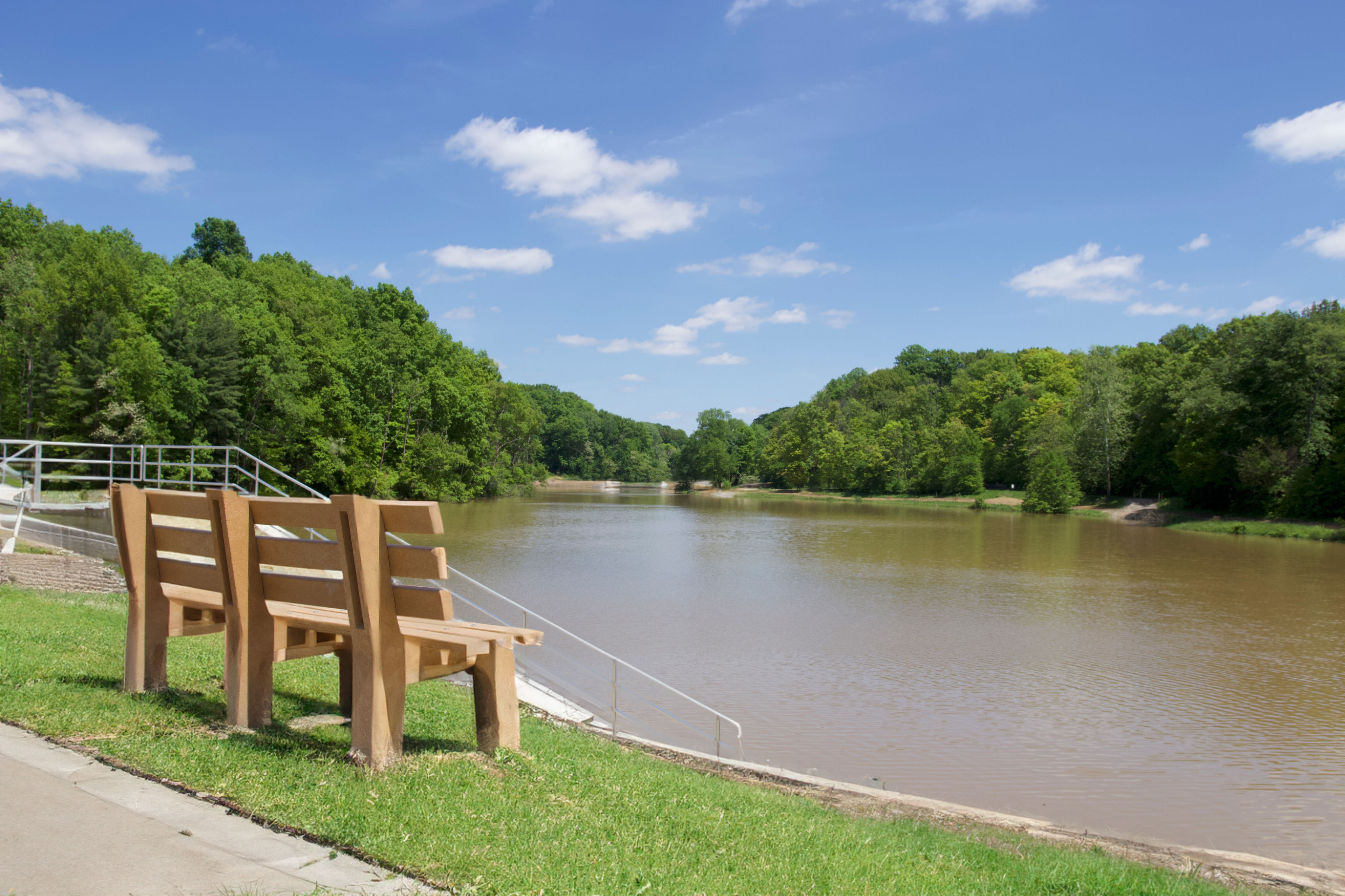 a view of a park bench facing mt gilead lake on a blue sky day with full green trees surrounding