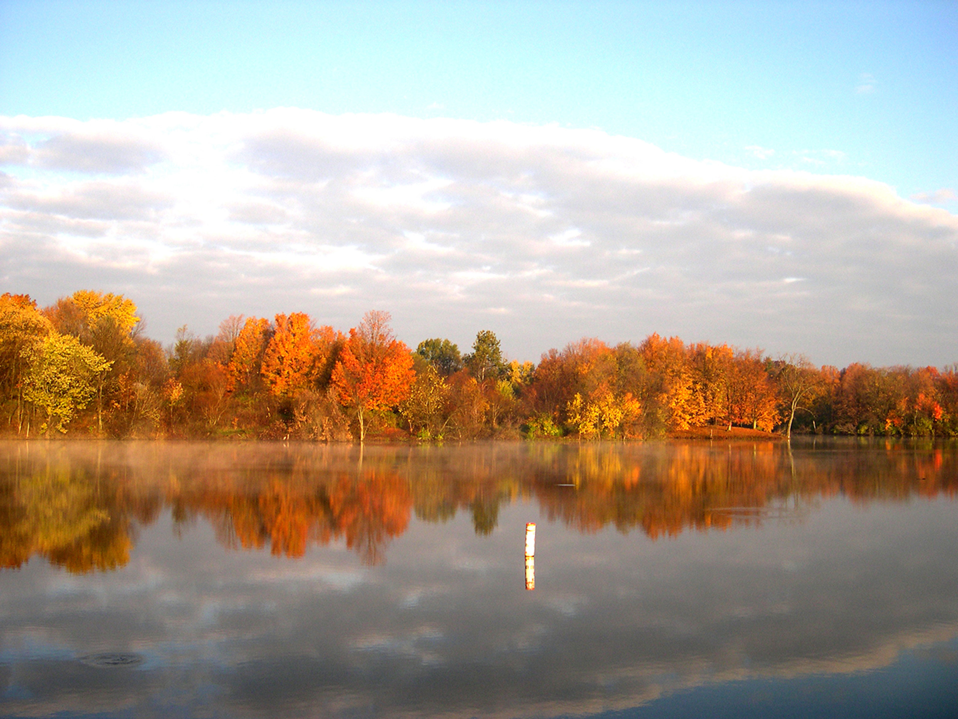 A body of water with orange and red trees and clouds in the sky at Madison Lake State Park