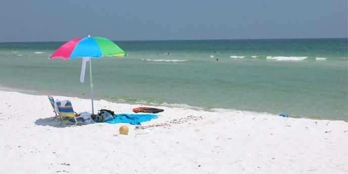 A beach umbrella shades a chair as blue-green waves crash against a white sand beach.
